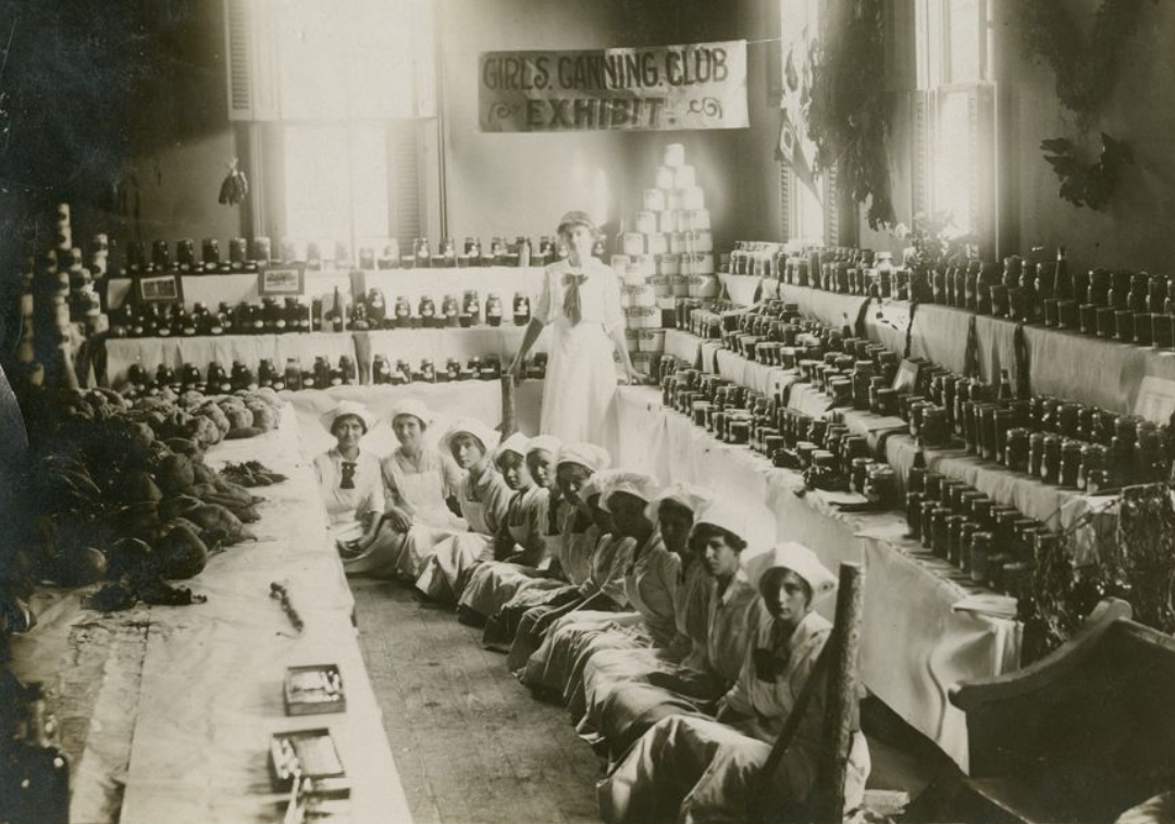Black and white photograph of a group of young women sitting on the floor, surrounded by shelves full of jars and cans