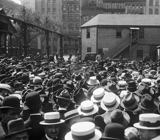 Black and white photograph of Emma Goldman in distance, surrounded by crowd of people wearing hats