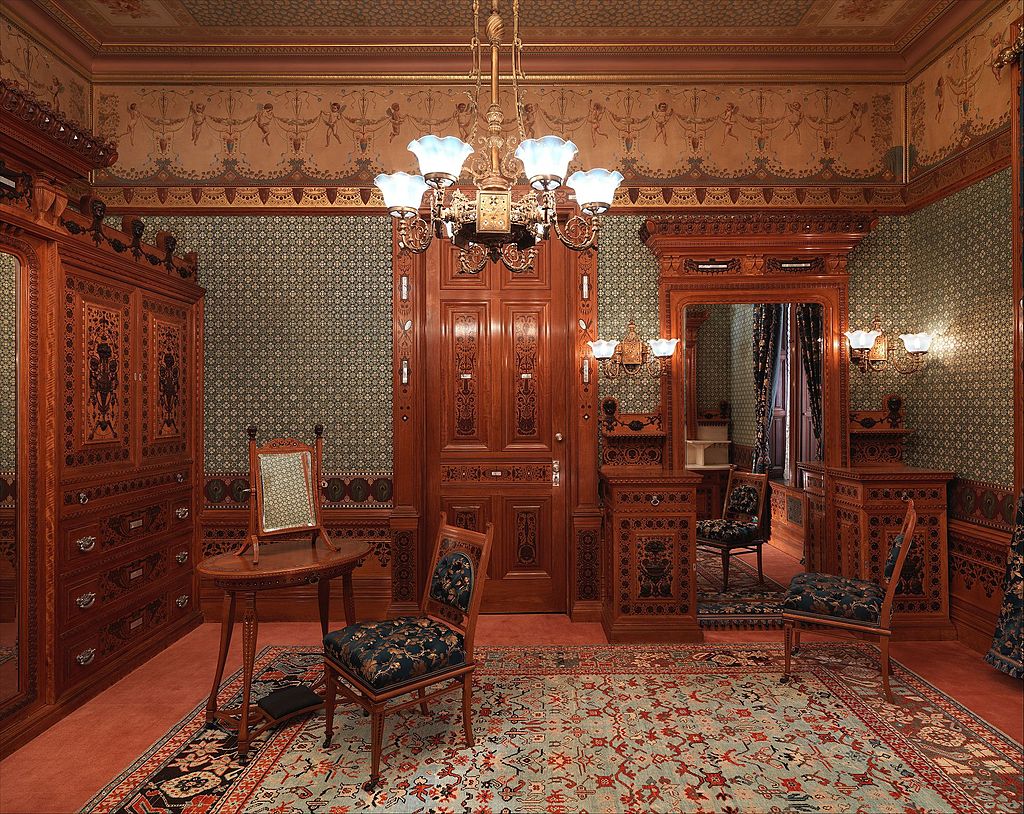 Photograph of a room featuring intricate wood paneling, green patterned wallpaper, wood furniture, and a chandelier