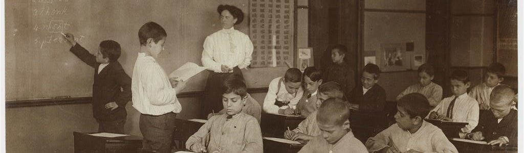 Image of teacher standing behind school children writing at their desks and on the board