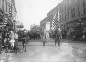 Photograph of a masked boy riding a bicycle next to flag bearers leading WWI victory parade