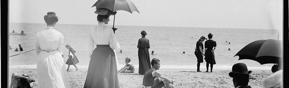 Image of people standing and sitting on a beach, some holding umbrellas.