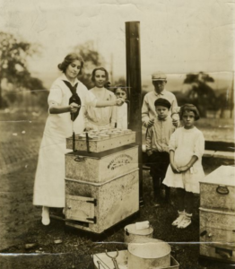 Black and white photograph of young woman standing with collection of cans, with five children standing around
