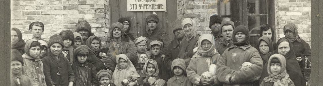Black and white photograph of crowd of men, women, and children gather in front of a soup kitchen
