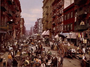 Colorized photograph of a crowded street, with carriages, pedestrians, and market stalls filled with fruits and vegetables