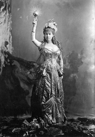 Black and white photograph of a woman wearing an elaborate dress, holding a glowing torch above her head